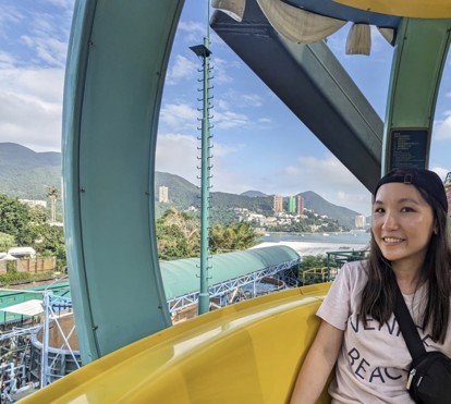 An Asian woman in a white t-shirt and black backwards hat smiles inside a Ferris wheel in Hong Kong.