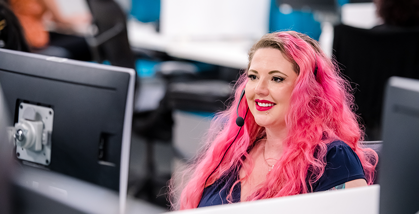 A woman with pink hair and wearing a headset sitting at a desk in an office, focused on her work.