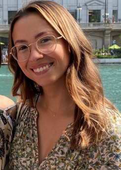 A young woman wearing glasses and a floral top smiles in front of a river.