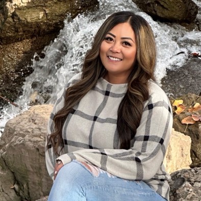 An Asian woman wearing a white and black plaid sweater smiles in front of a rushing stream.