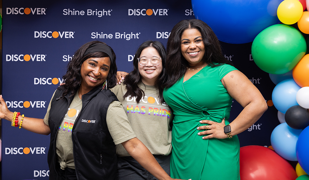 Three employees smiling in front of a Discover-branded backdrop with balloons 